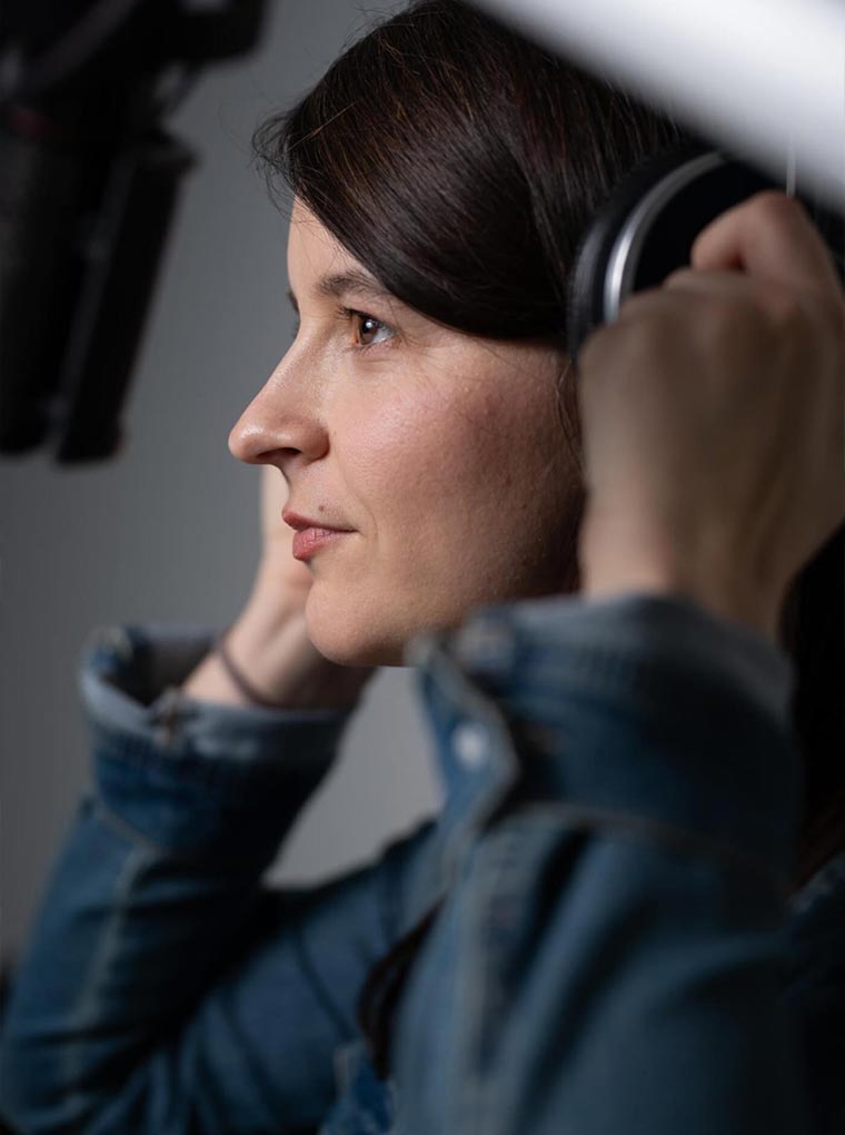 Marlene Groihofer von audioportait interviewt einen Holzarbeiter mit Motersäge im Freien. Im Hintergrund sieht man schöne Landschaft mit Wiesen, Hügeln und schneebedeckte Bäume. 