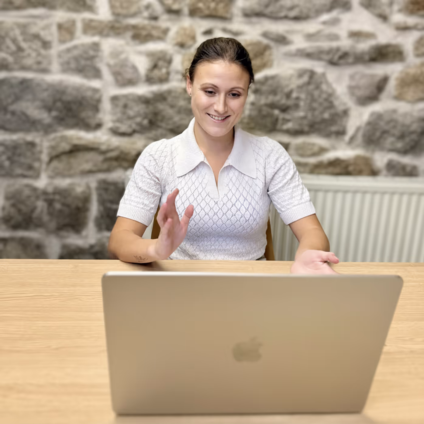 Frau (Jennifer) mit weißem T-shirt sitzt vor einem Laptop und lächelt, Steinwand im Hintergrund.