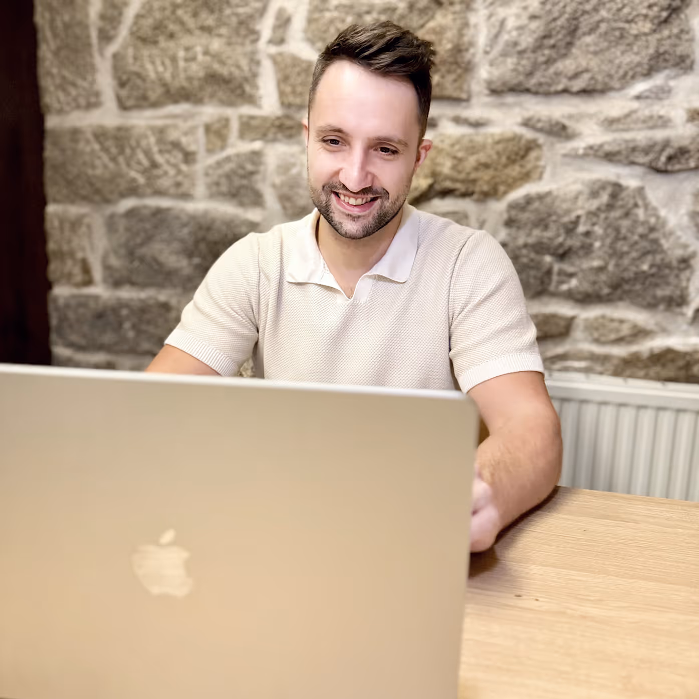 Ein lächelnder Mann (Timo) sitzt an einem Holztisch und arbeitet an einem MacBook vor einer Steinwand.