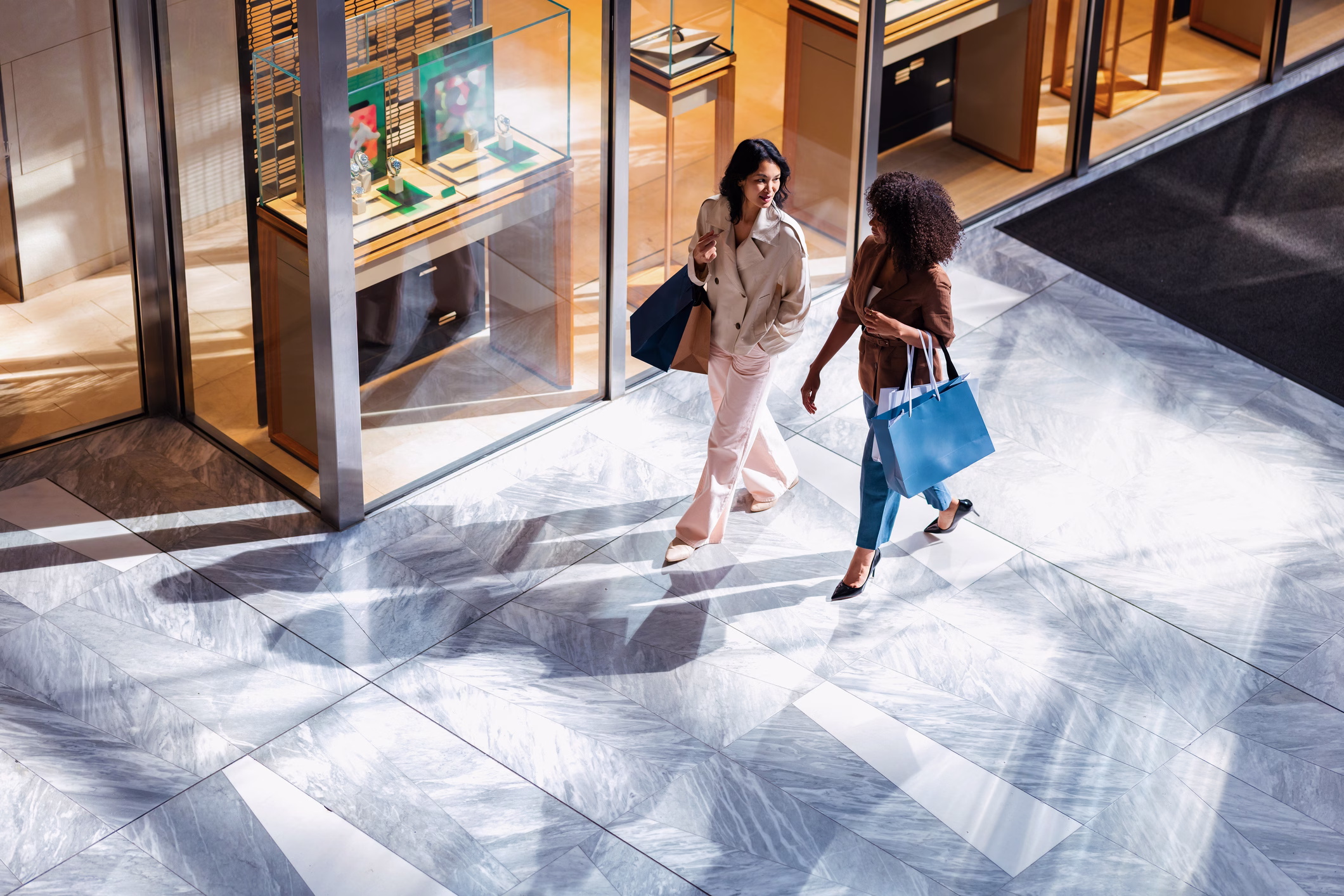 Two shoppers with bags crossing the sunlit marble concourse of a luxury retail gallery