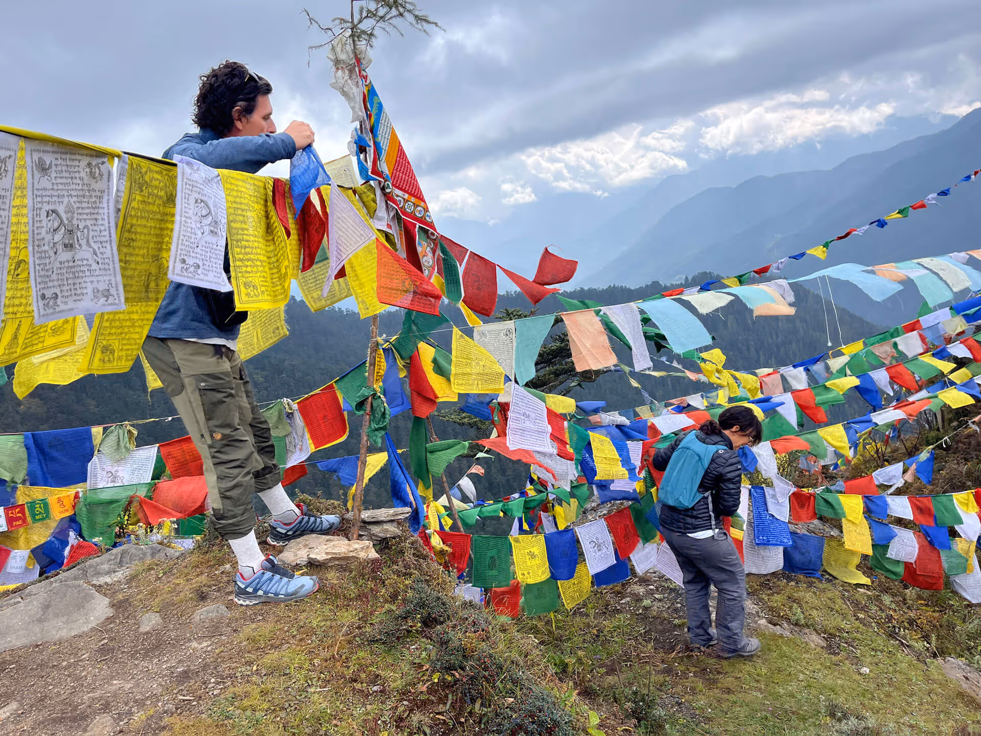 Traveler tying colorful prayer flags on a mountain pass in Bhutan, with misty Himalayan peaks in the background.