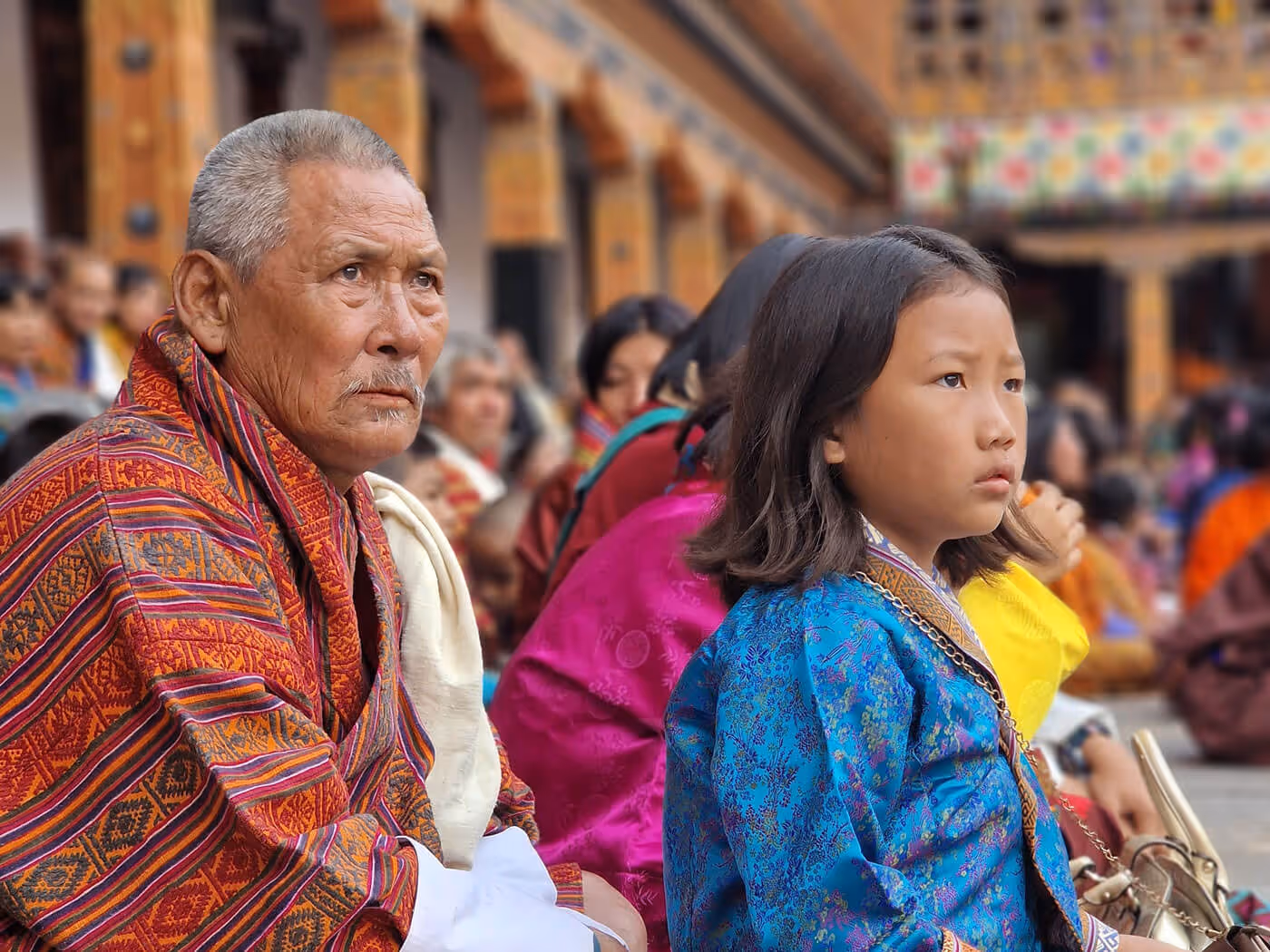 Elderly Bhutanese man and young girl in traditional dress watching festival, embodying generational continuity.
