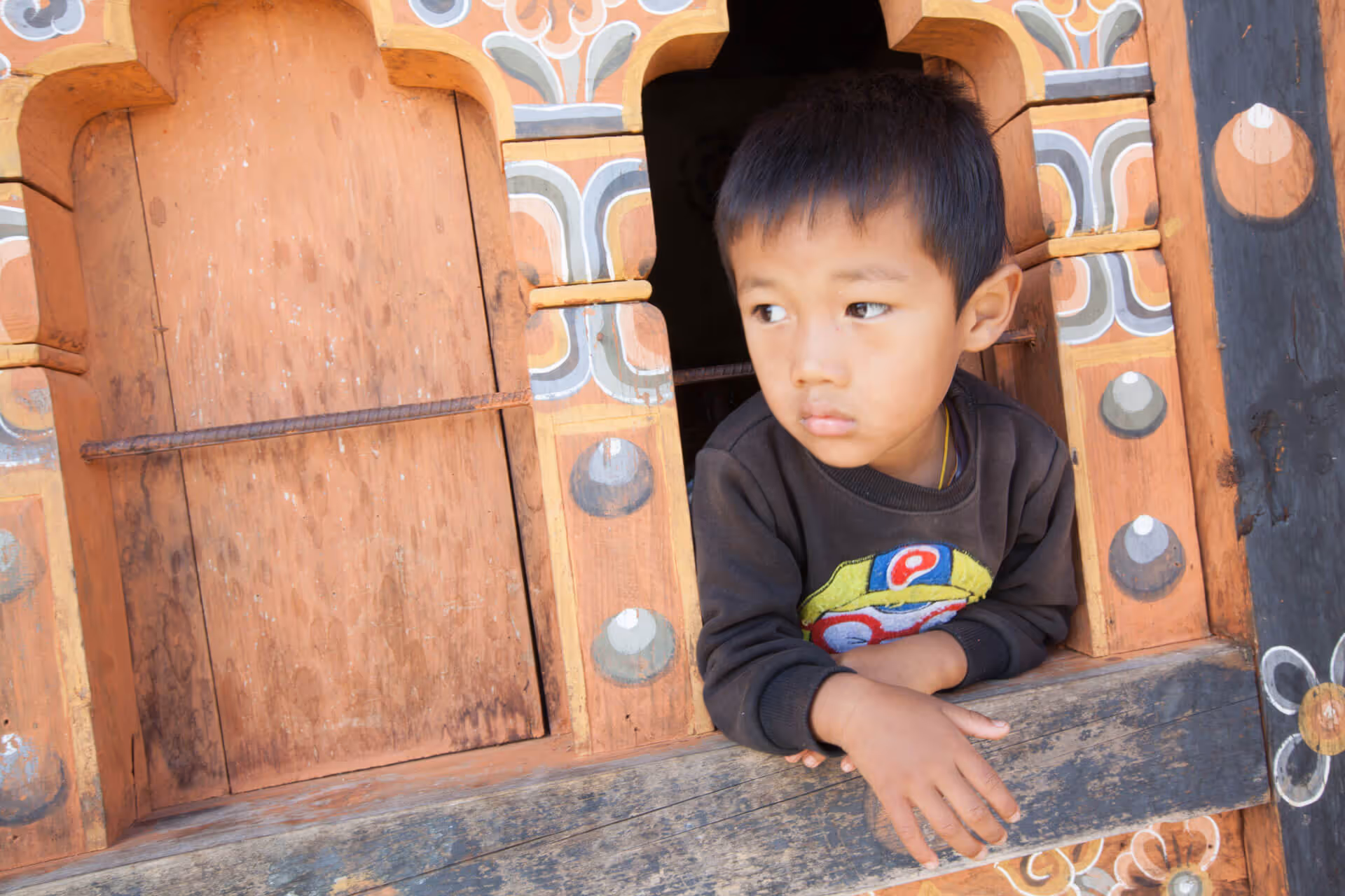 Young Bhutanese boy gazes out from a traditional wooden window, symbolizing hope for future generations and Raven Tours' purpose-driven travel values.
