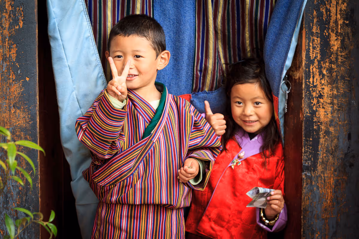 Two Bhutanese children in traditional dress smile warmly at the camera, flashing peace signs and thumbs-up outside a doorway.