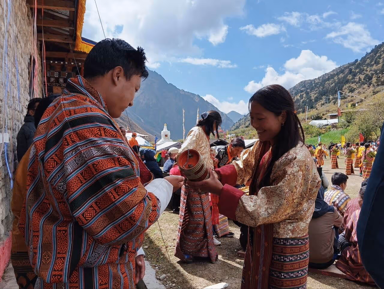A Bhutanese woman in kira offers a ceremonial drink to a guest during a mountain festival, celebrating tradition, hospitality, and shared joy.