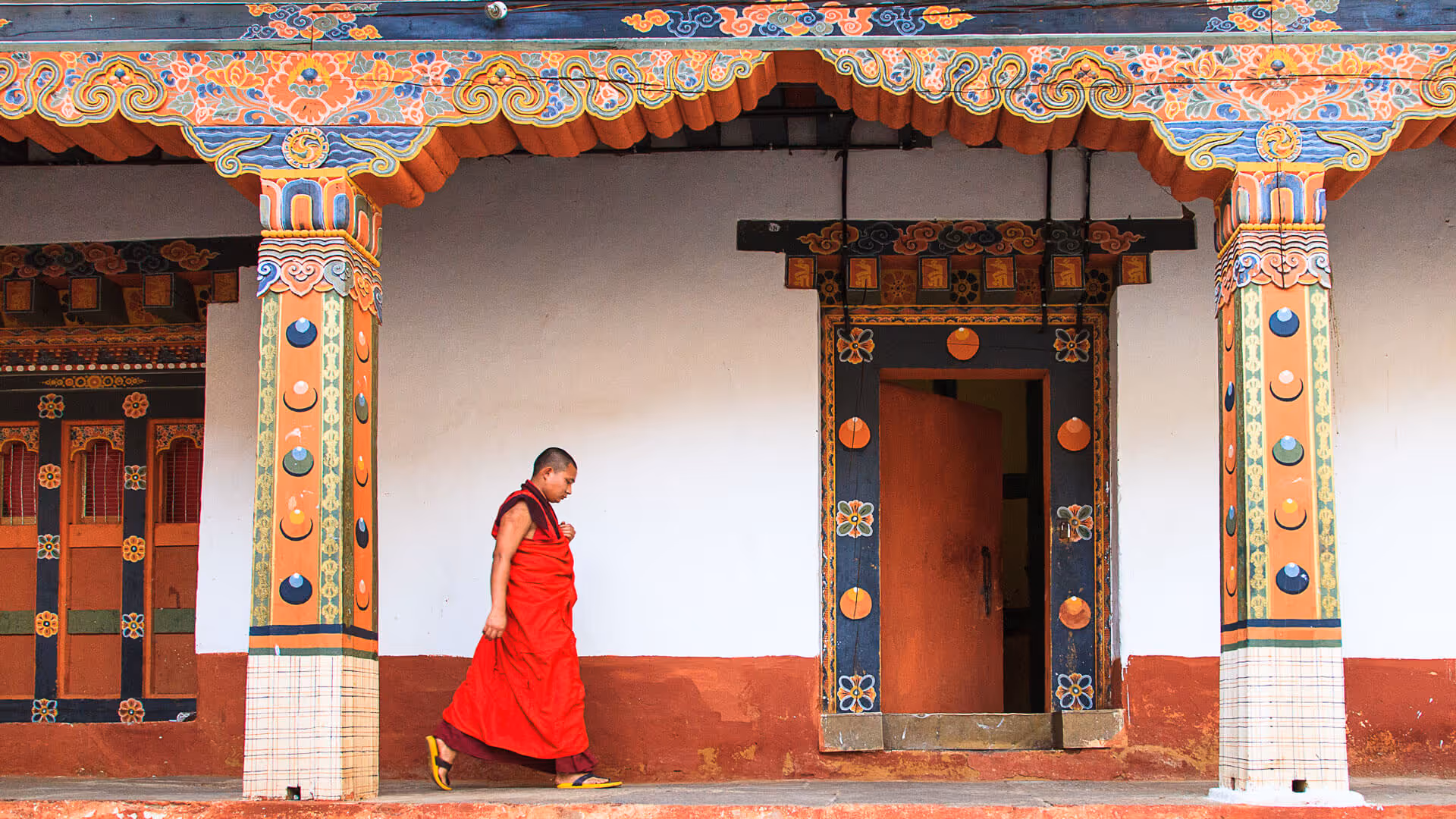 A monk in red robes walking slowly past vibrantly decorated columns and colorful wooden doorways at a Bhutanese monastery.