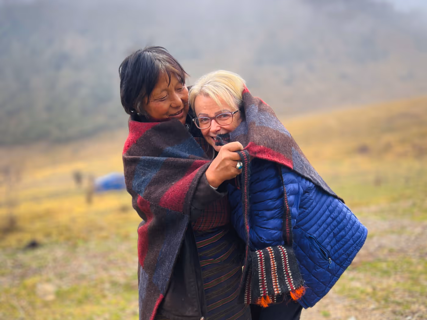 A Raven guest and local friend wrapped in a shared wool blanket, sharing laughter in Bhutan’s misty highlands.