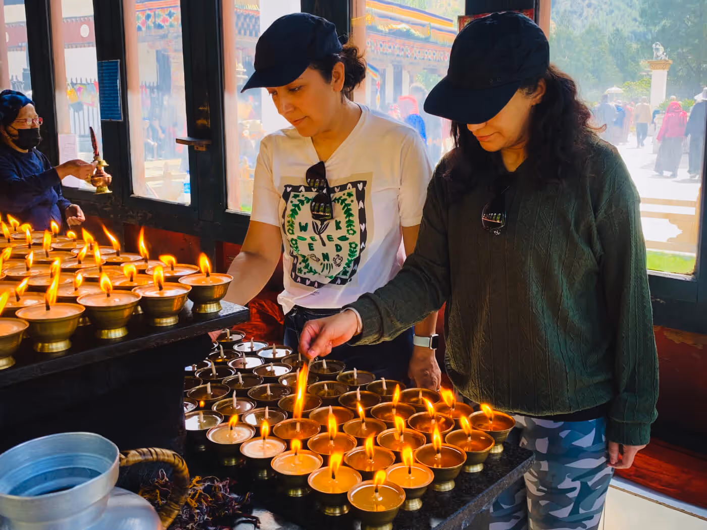 Two travelers light butter lamps in a serene monastery courtyard, participating in Bhutan’s timeless act of prayer and devotion.