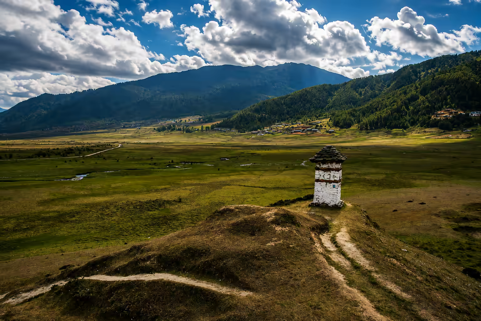 A quiet Bhutanese valley with a lone chorten, winding paths and forested mountains beneath dramatic clouds.