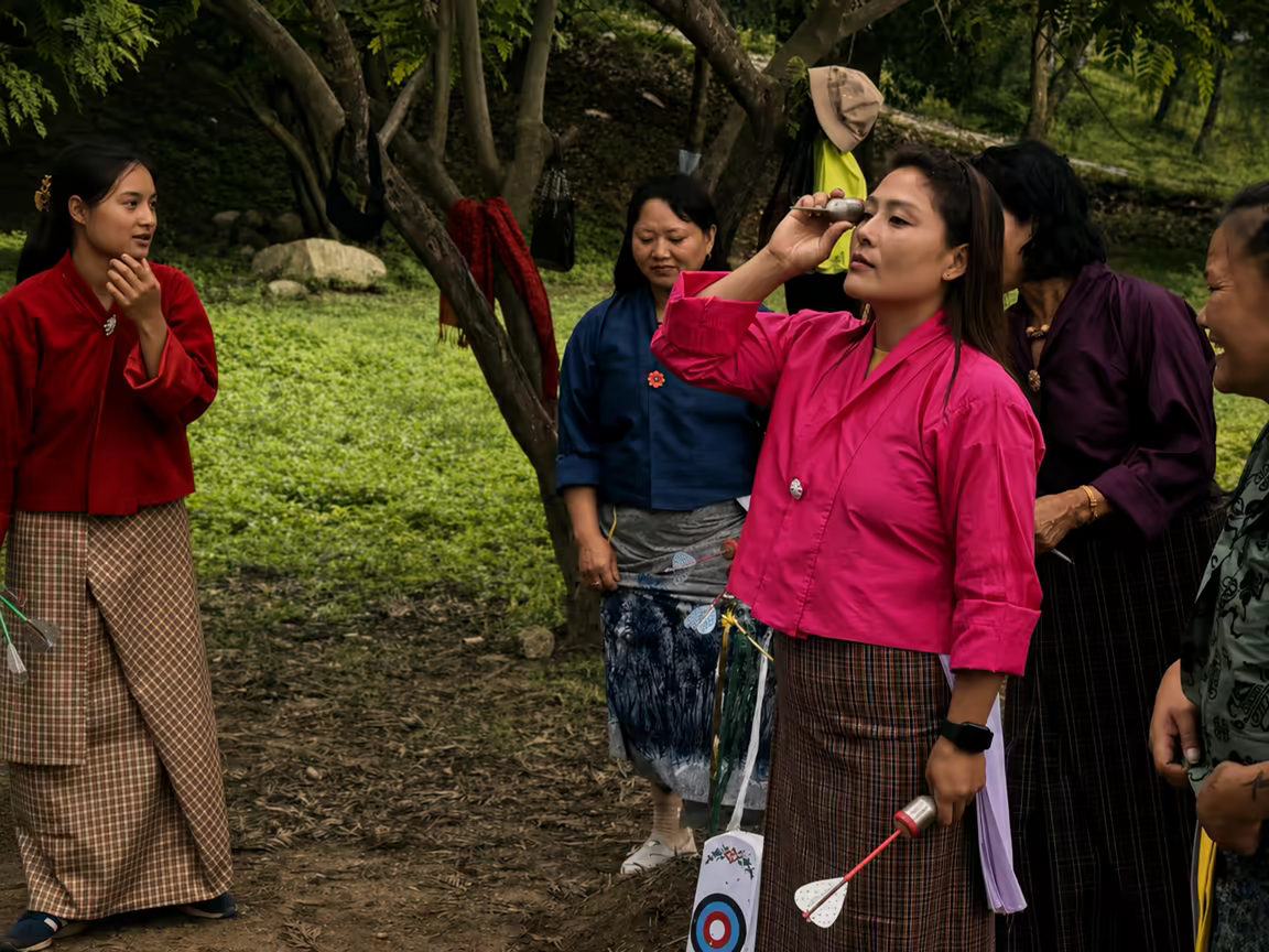 Bhutanese women play khuru, a traditional dart game, beneath forest trees.