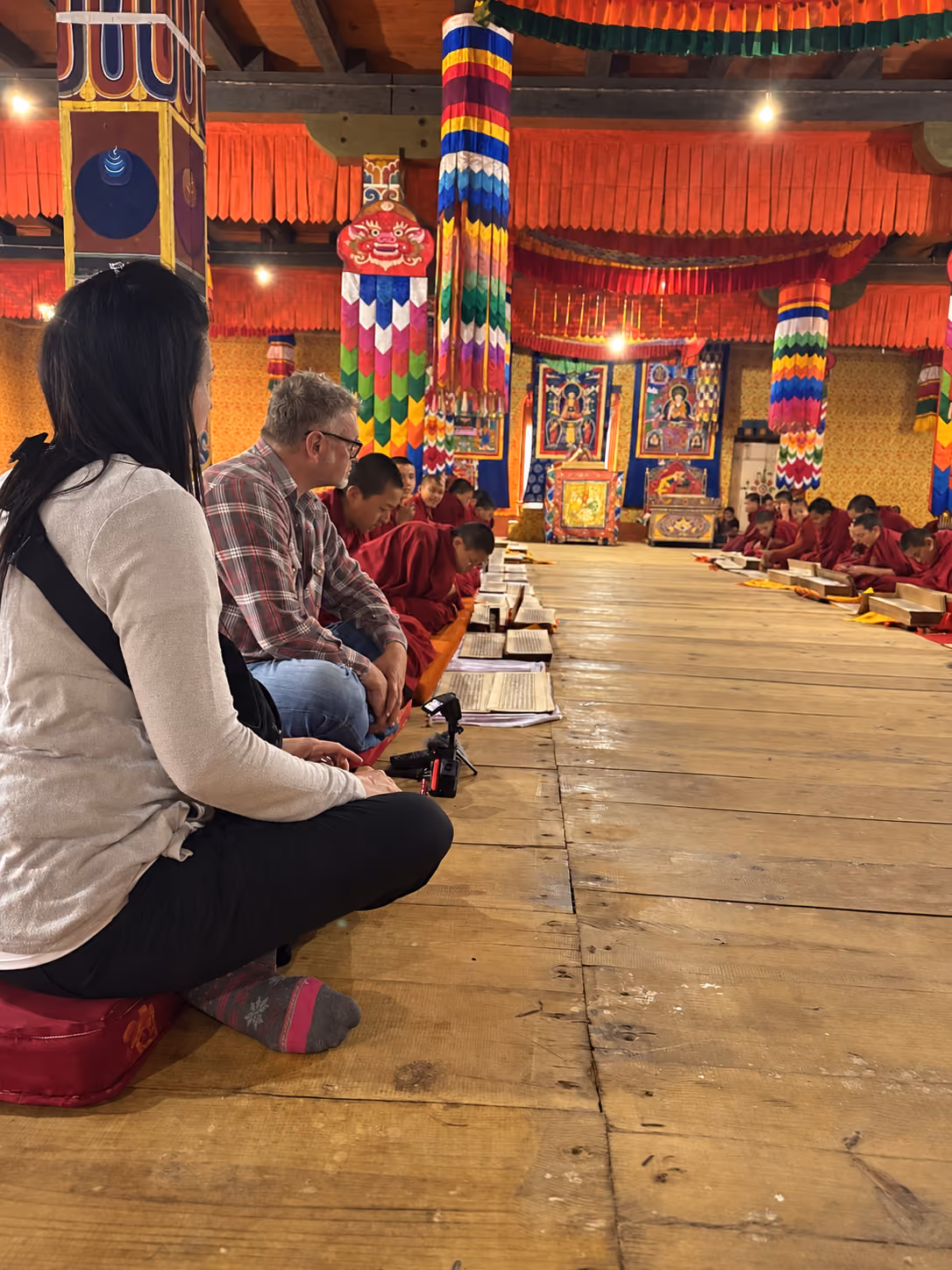 Visitors sit beside monks reading scriptures in a richly decorated Bhutanese prayer hall.