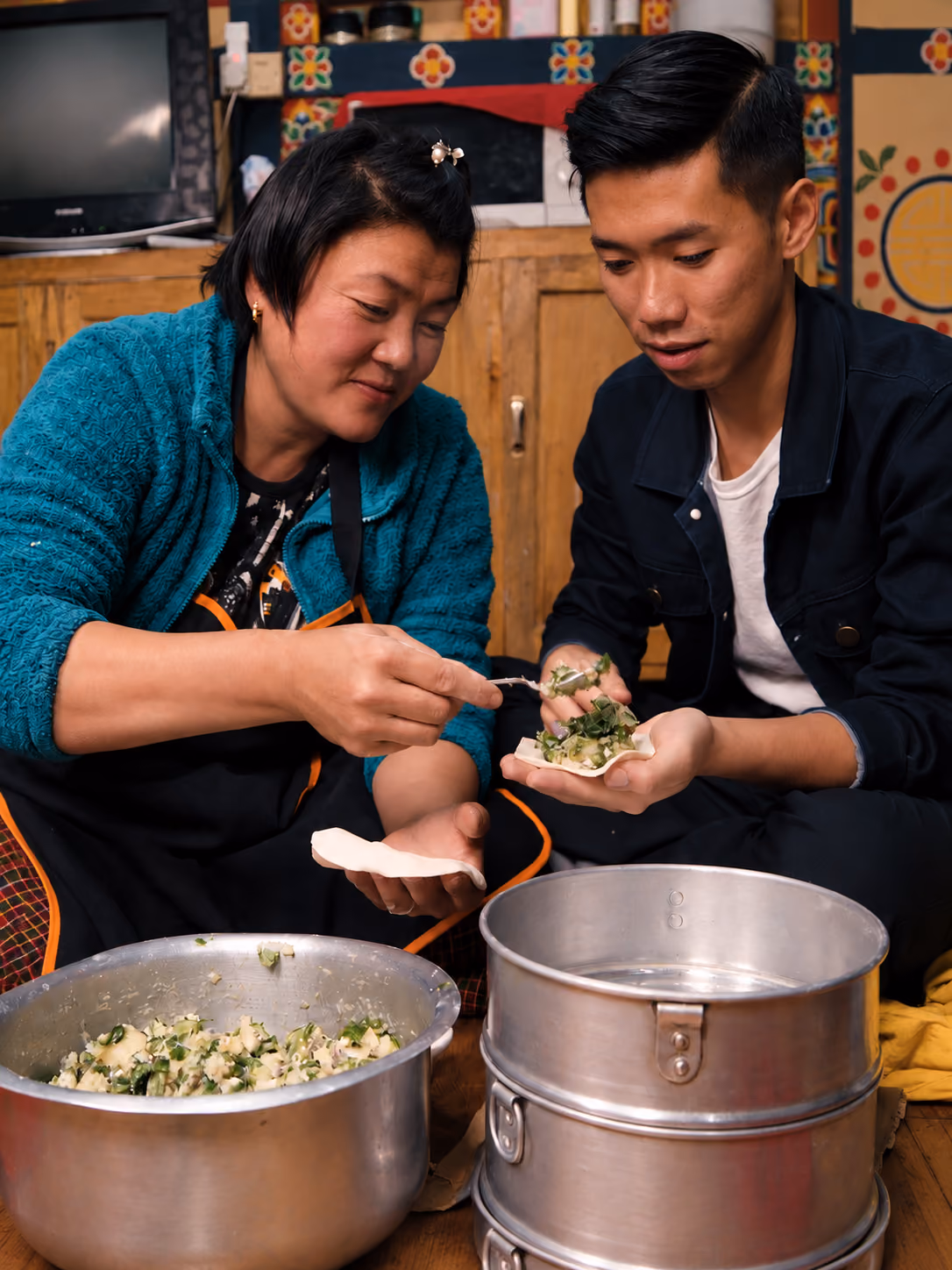 Two people prepare momos together, spooning vegetable filling into dough in a Bhutanese kitchen.