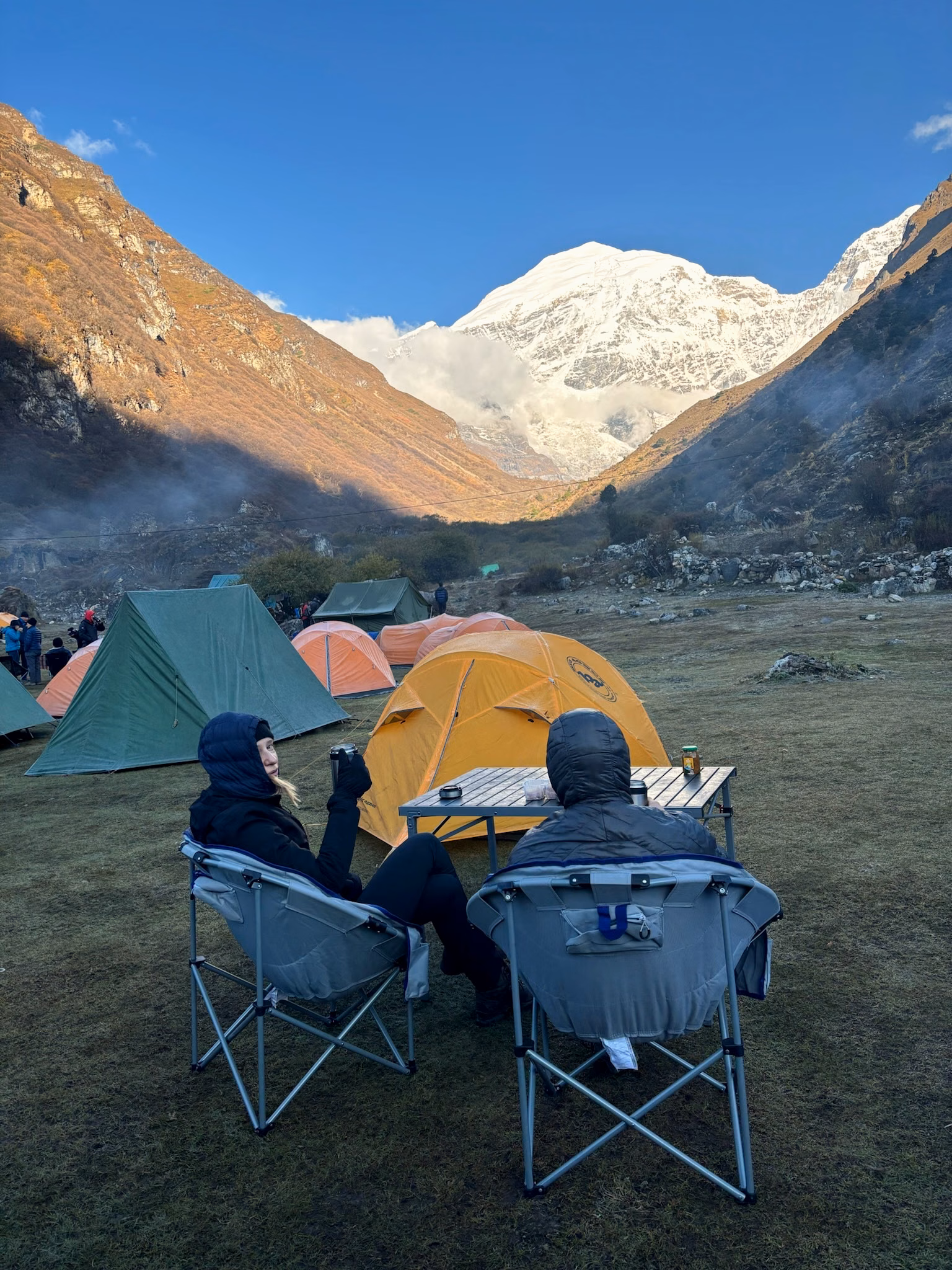 Two guests relaxing beside orange tents at a supported high-altitude camp beneath Jomolhari in Bhutan.