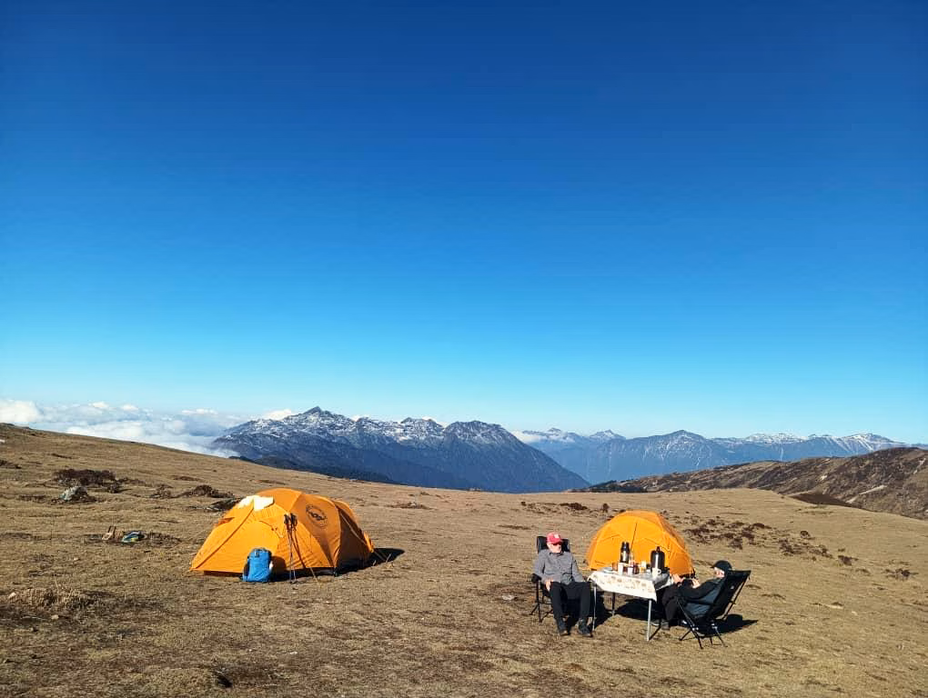 Two guests seated beside orange trekking tents at a supported mountain camp in Bhutan.