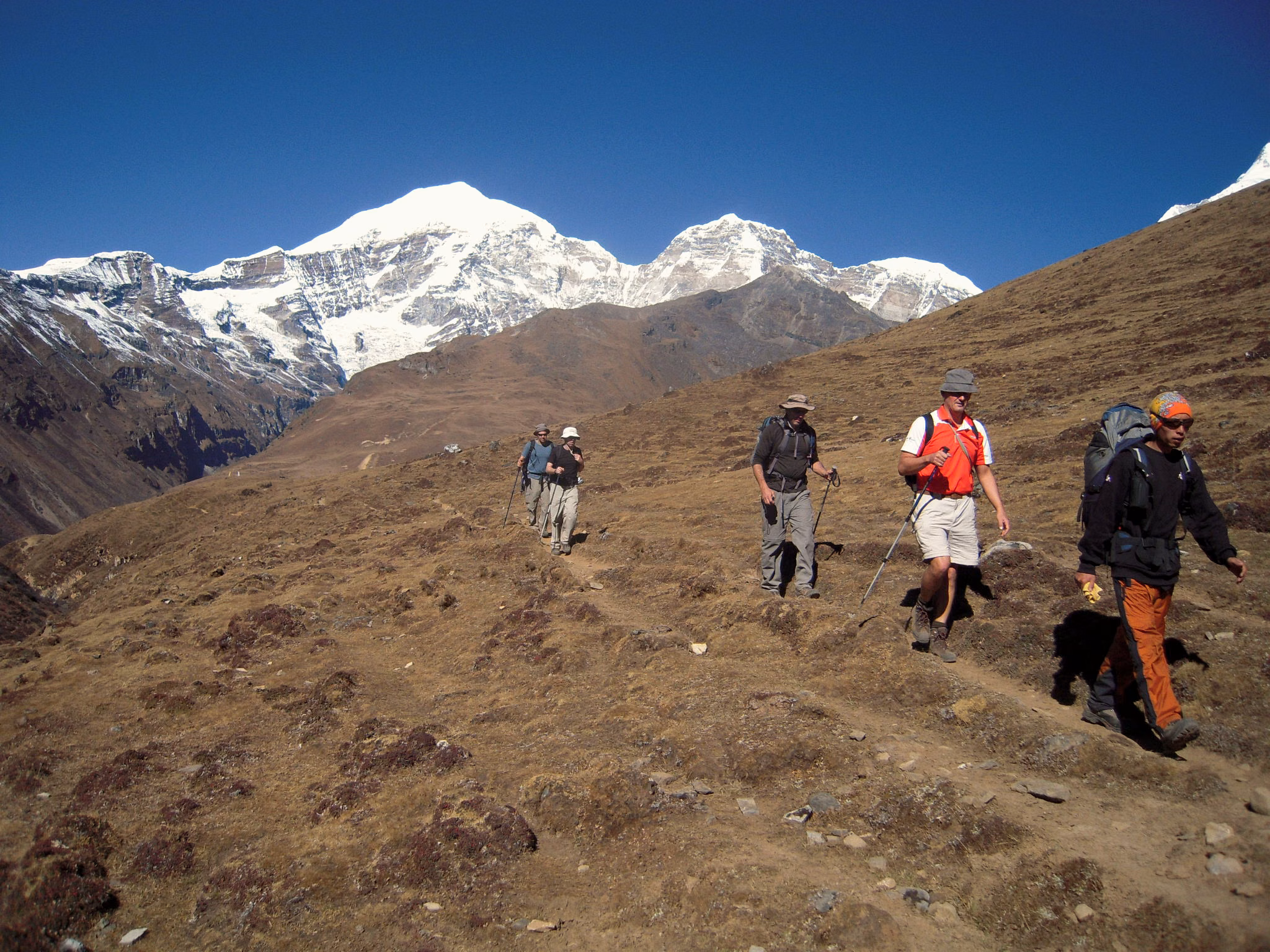 Trekkers walking across a high alpine slope beneath snow-covered peaks on the Jomolhari route in Bhutan.