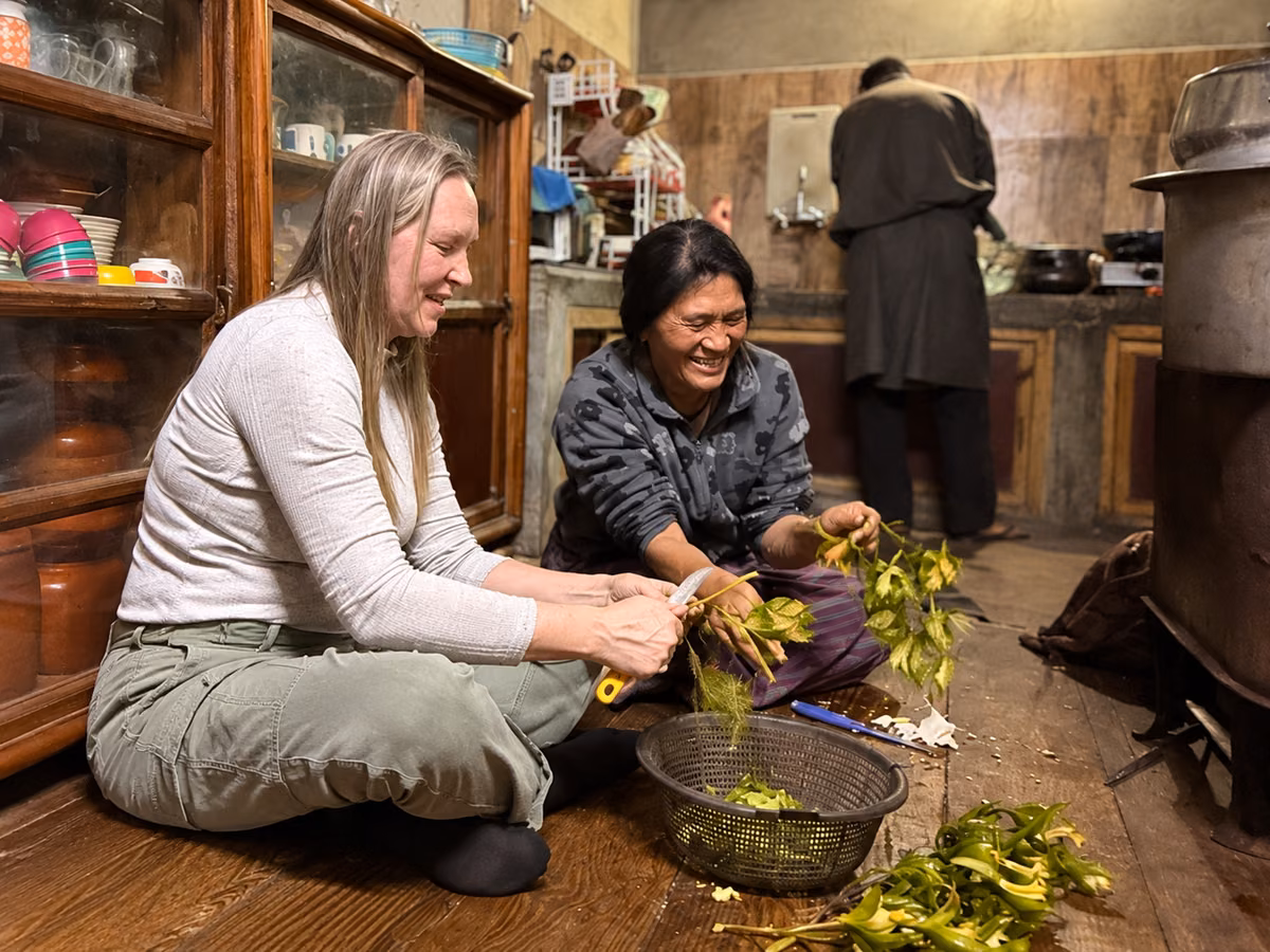 A guest and Bhutanese host smiling as they prepare vegetables in a farmhouse kitchen.