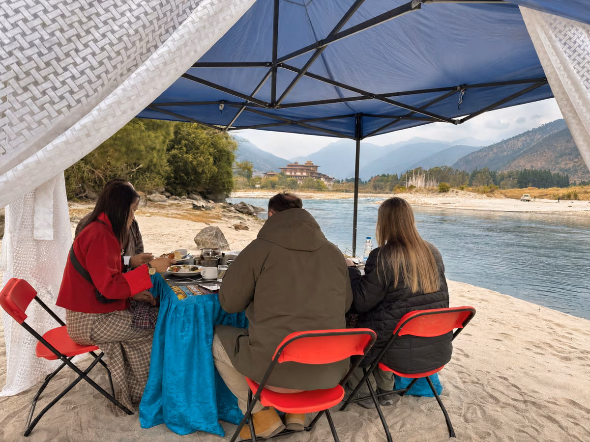 Guests sharing lunch beneath a canopy beside the river, with Punakha Dzong in the distance.