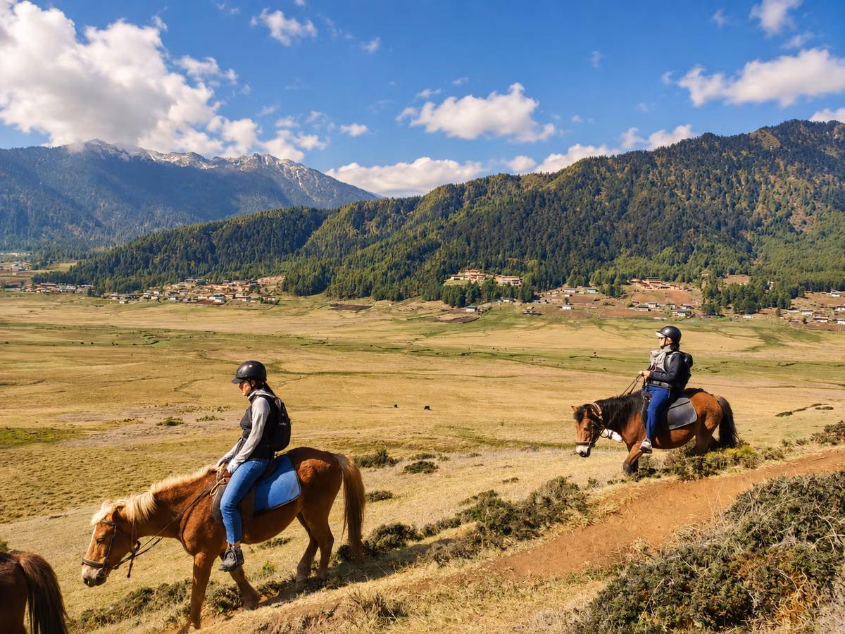 Two helmeted guests riding horses along a hillside trail above a broad Bhutanese valley.