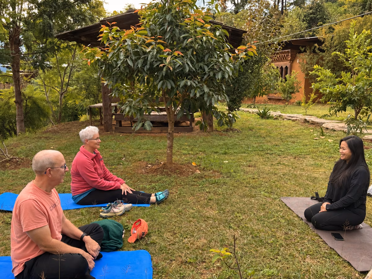 Two guests seated on yoga mats with an instructor in a Bhutanese garden.