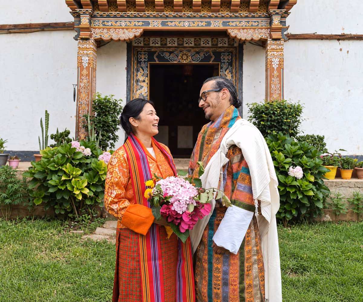 A laughing couple with a bouquet and ceremonial scarves outside an ornate Bhutanese doorway.