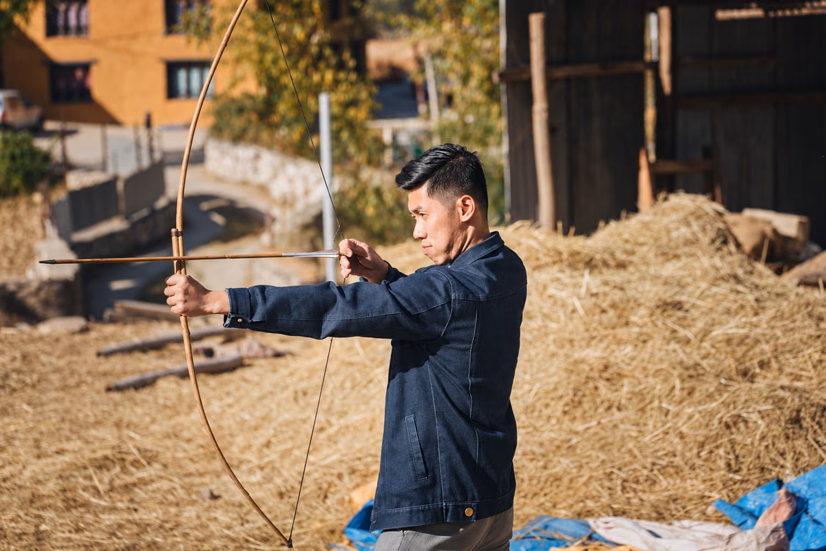 An archer drawing a traditional bamboo bow beside a rural Bhutanese building.