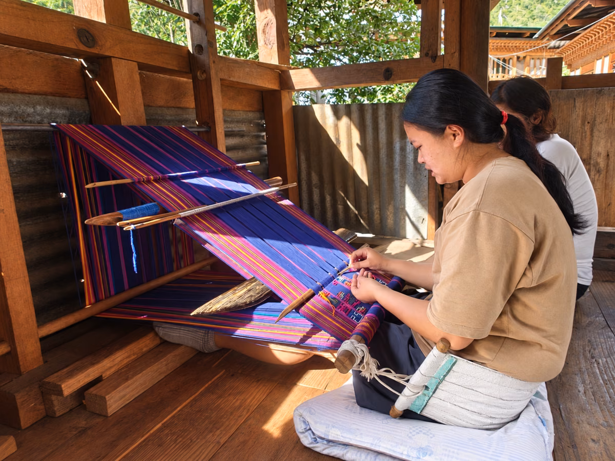 A woman weaving colourful patterned cloth on a traditional backstrap loom in a wooden workshop.