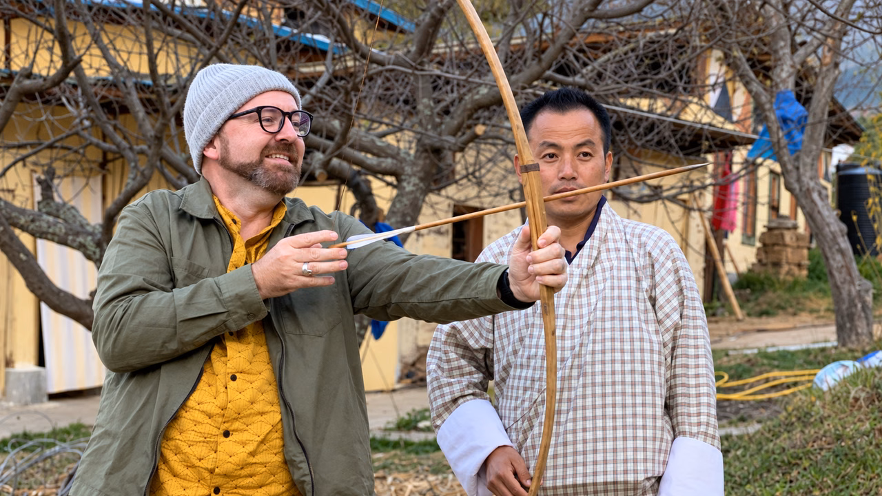 Guest trying a bamboo bow beside a Bhutanese man in traditional dress.