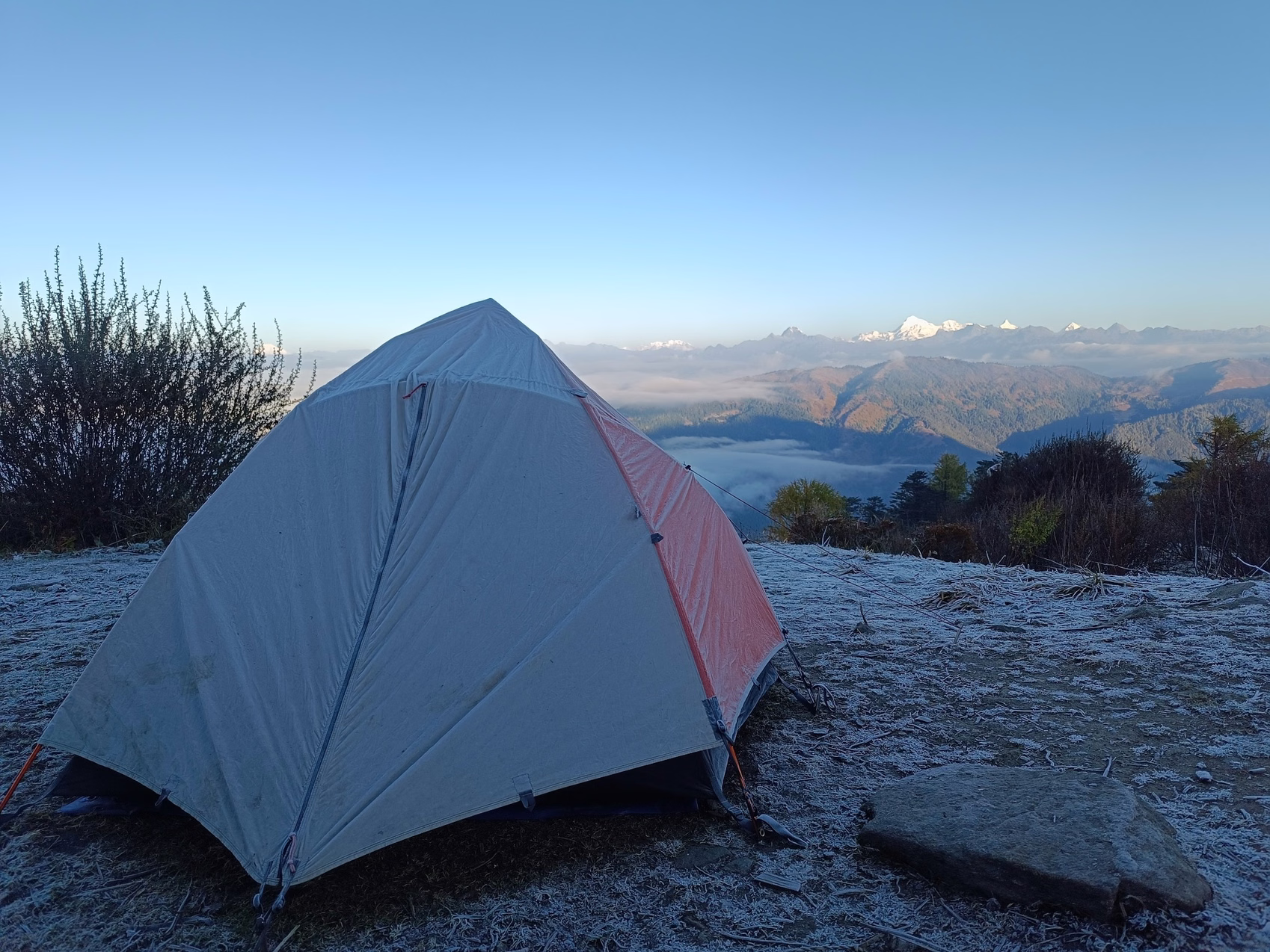 A frost-covered trekking tent overlooks mountain ridges and low clouds at sunrise in Bhutan.