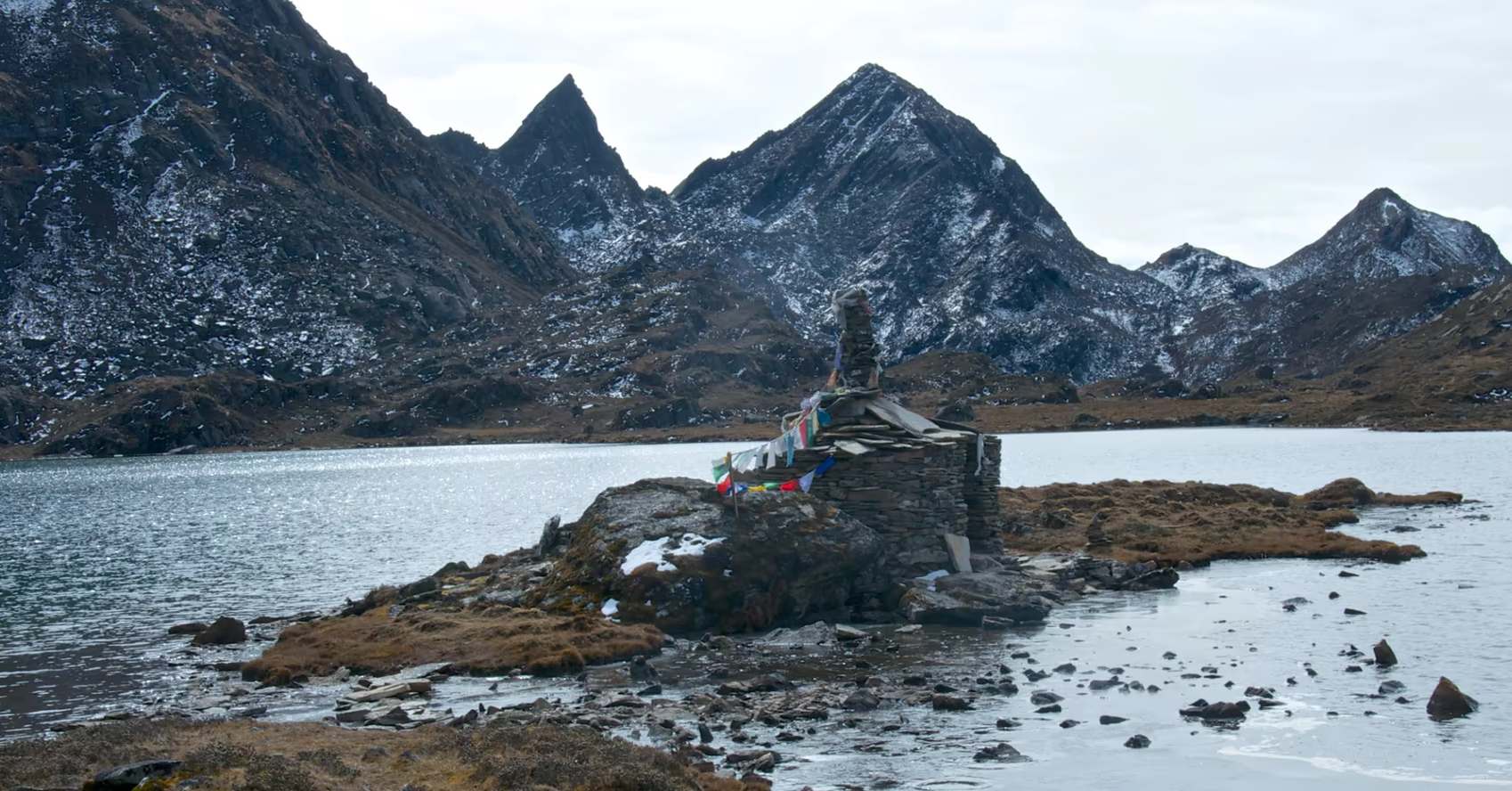 Stone shrine and prayer flags beside a Dagala alpine lake beneath snow-dusted ridges.