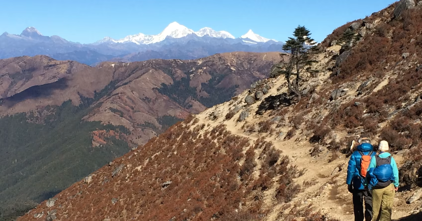 Two trekkers following a hillside trail in Dagala with distant snow-covered Himalayan peaks.