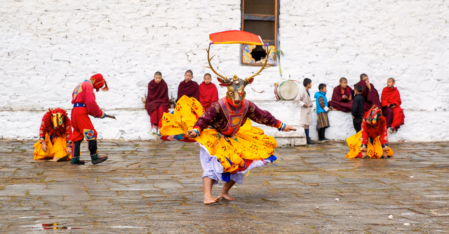 Masked dancer performing at Paro Tshechu in Bhutan, with monks watching beside the courtyard wall