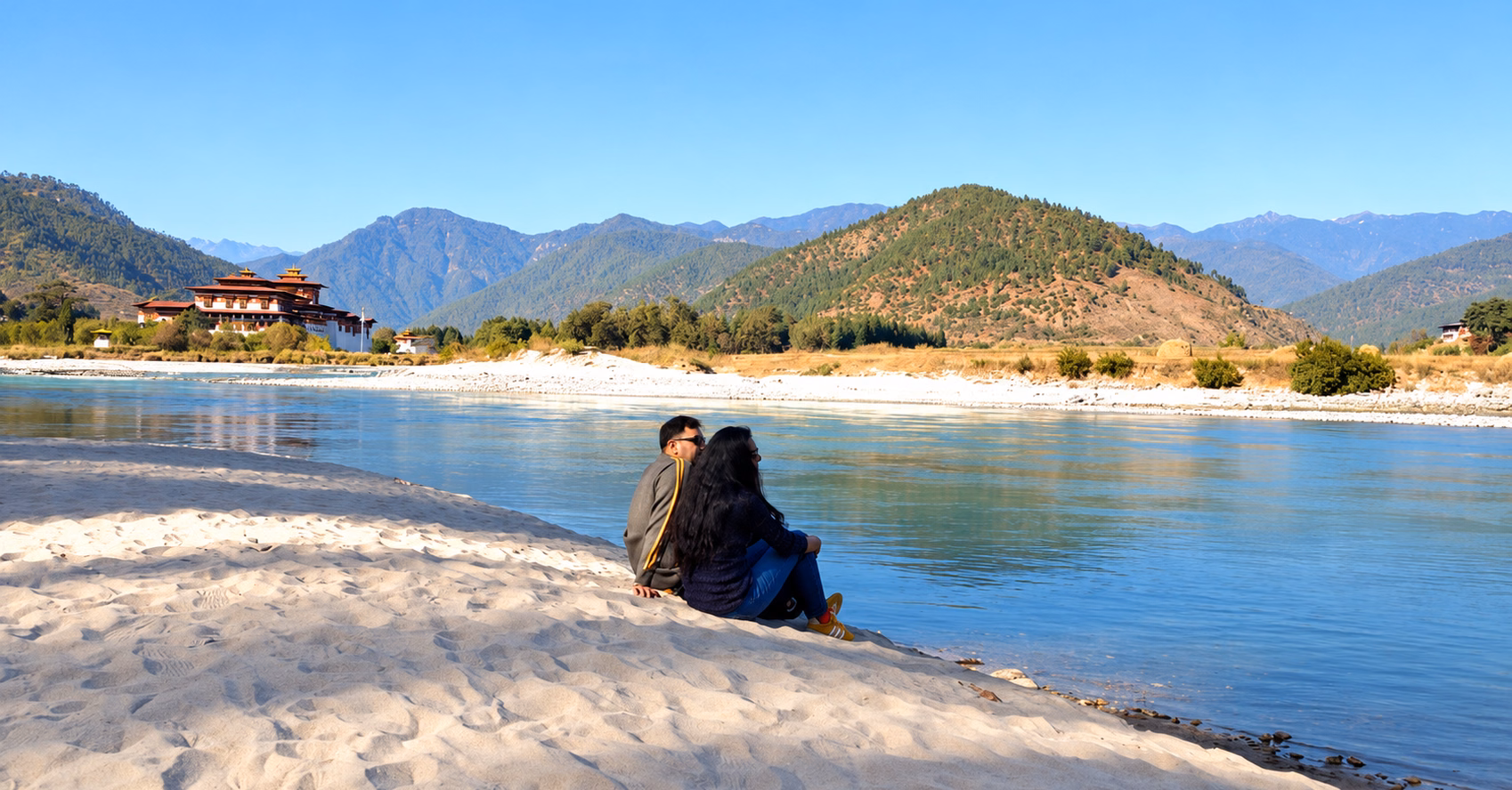 Two guests relaxing beside the river with Punakha Dzong and mountains beyond in Bhutan