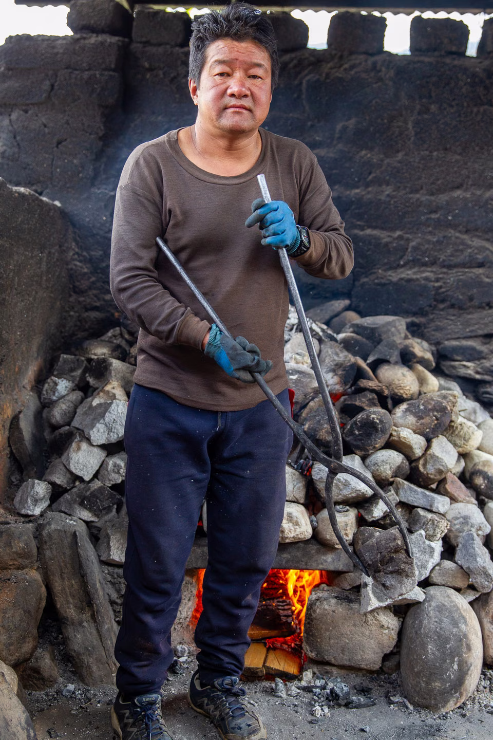 Hot-stone-bath attendant holding metal tongs beside stones heating over a fire.