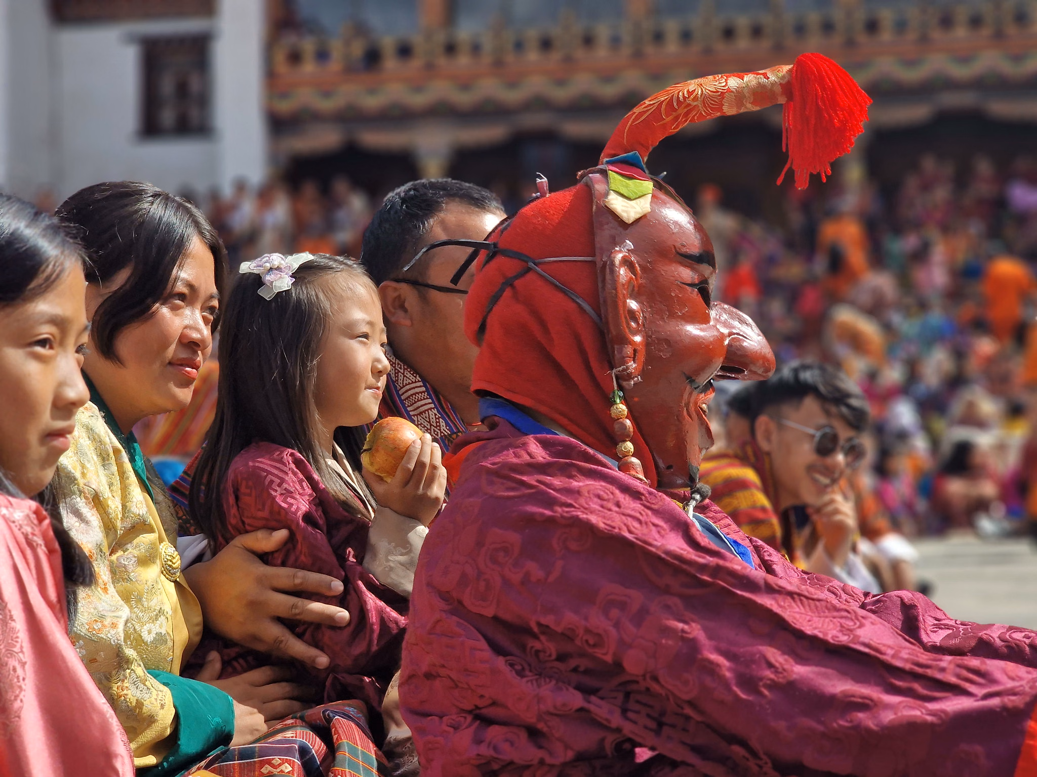 A red-masked performer seated beside festivalgoers, including a child holding an apple.