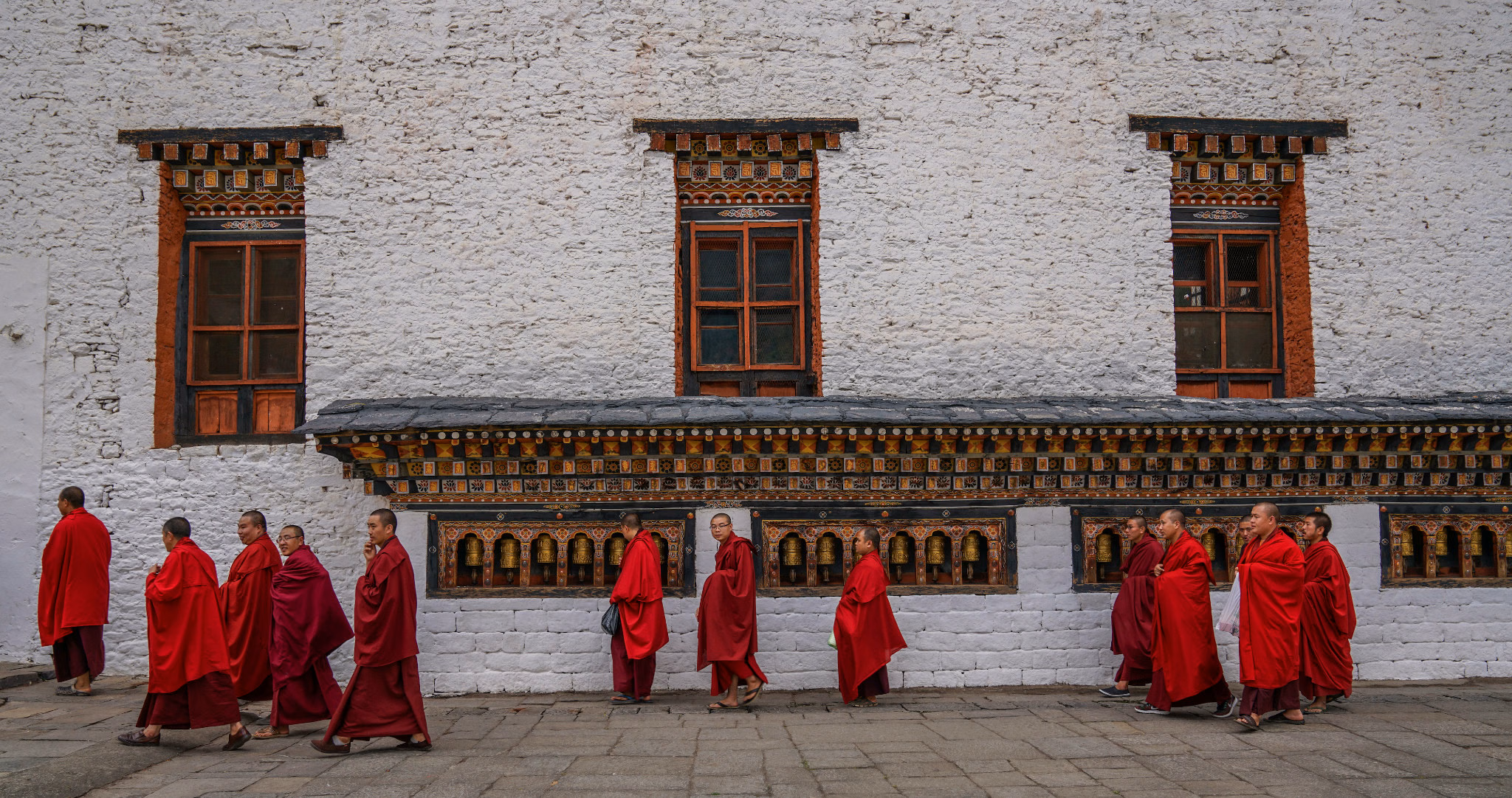 Monks in red robes walking beside a row of prayer wheels beneath a white wall.