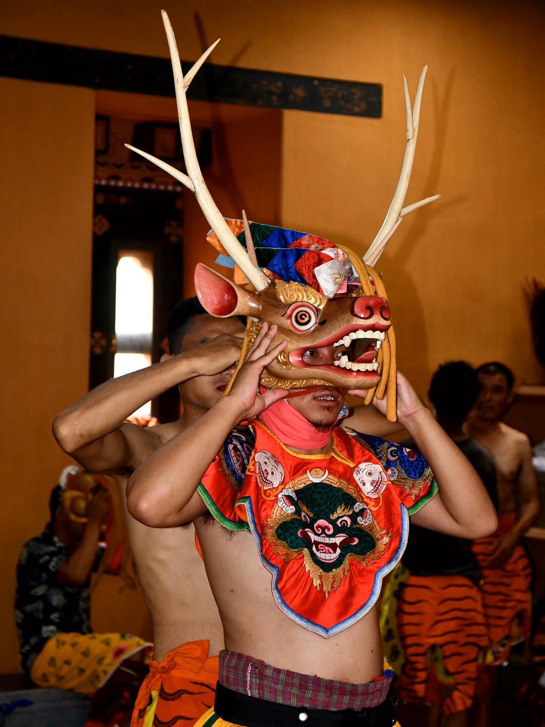 A performer holding an antlered mask above his face while adjusting it in an interior room.