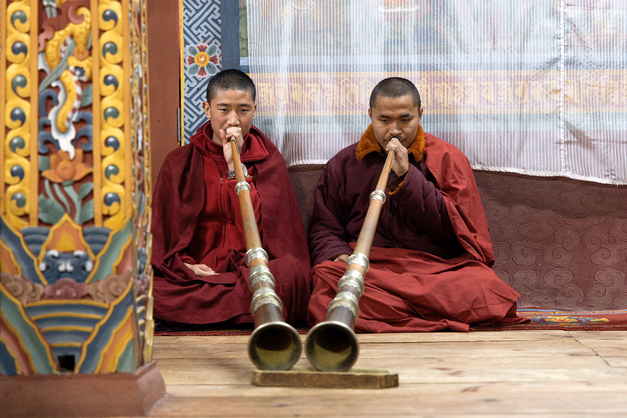Two monks seated in a decorated interior, playing long horns.