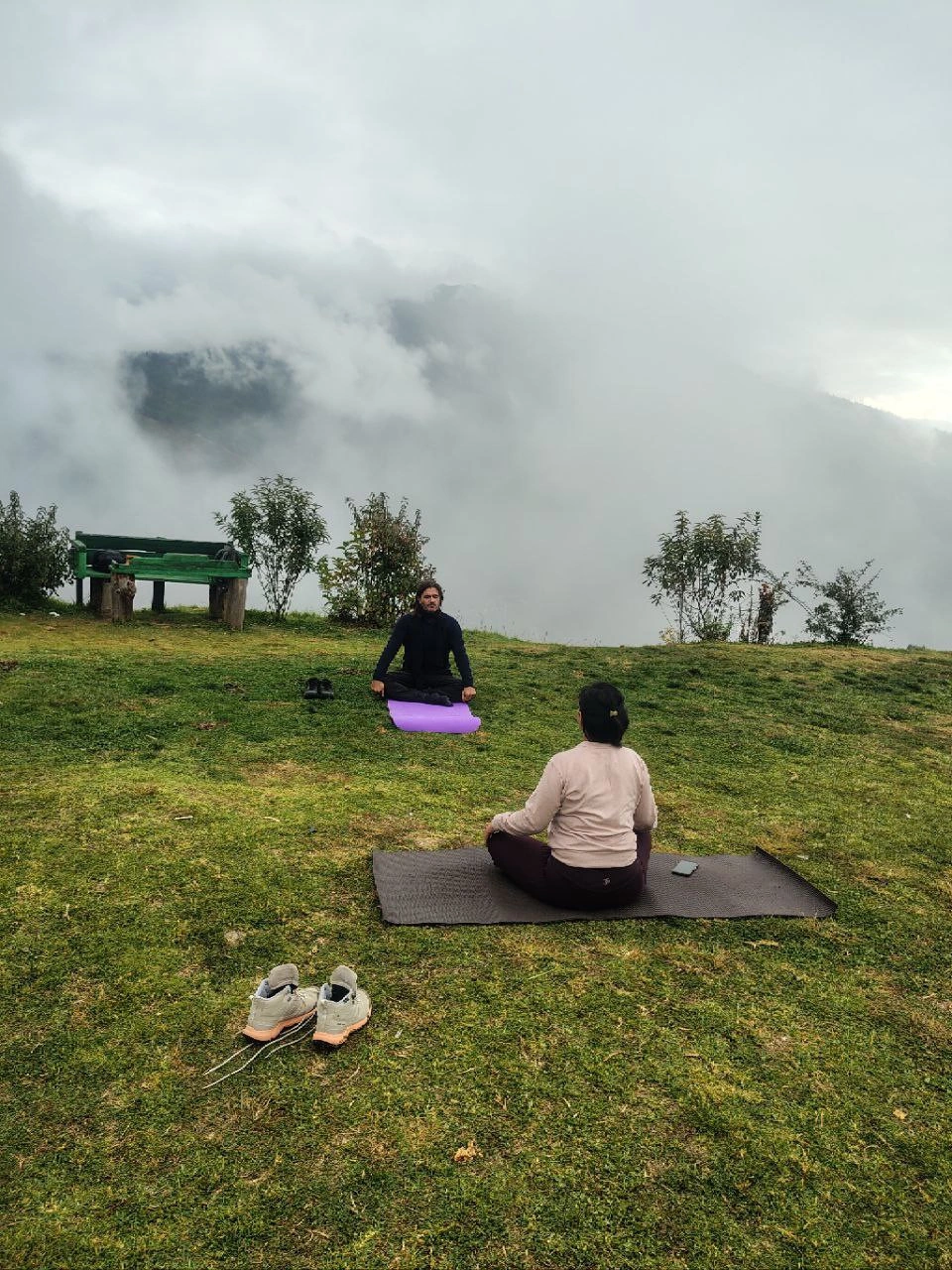 Two people seated on yoga mats on a grassy hillside in Bhutan, surrounded by mountain mist.
