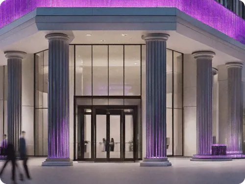 Modern building entrance with tall classical-style columns, large glass doors and windows, and purple accent lighting illuminating the columns and upper facade. People are walking near the entrance.