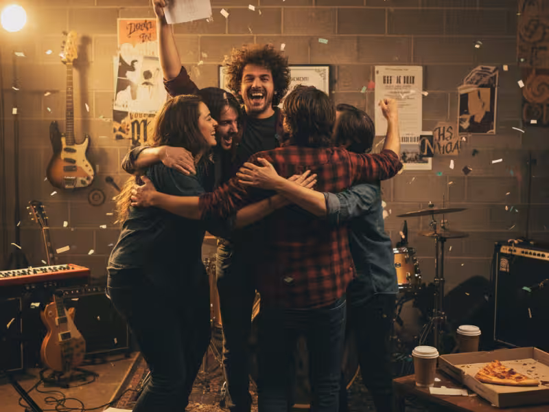 Band members hugging each other in a basement rehearsal space. One the band members has his hand up in the air with a letter, a congratulatory pose.
