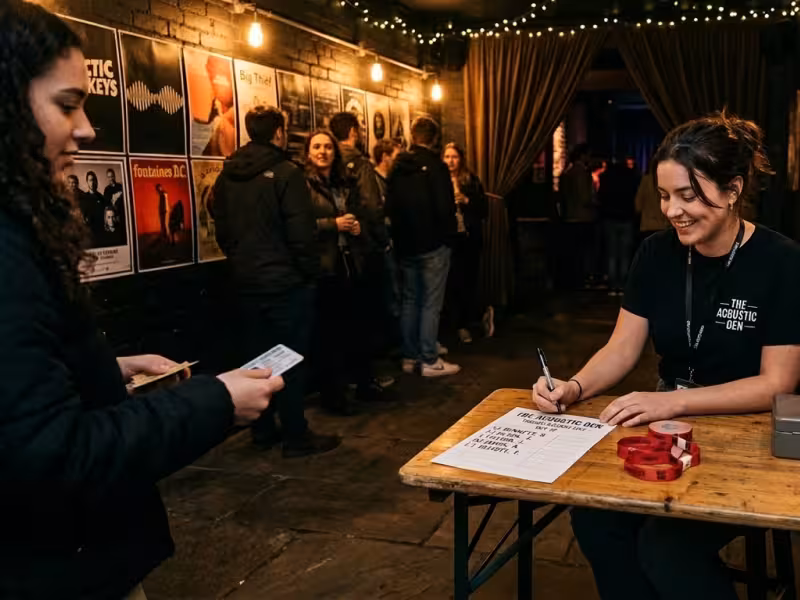 A photograph of a female staff member smiling while checking in attendees at a small, independent music venue. She sits behind a rustic wooden folding table featuring a printed guest list, a pen, a metal cash box, and red wristbands. The venue has a cozy, intimate atmosphere with dim, warm string lighting and dark brick walls covered in band posters. A small line of people waits in the background near a curtained entrance.