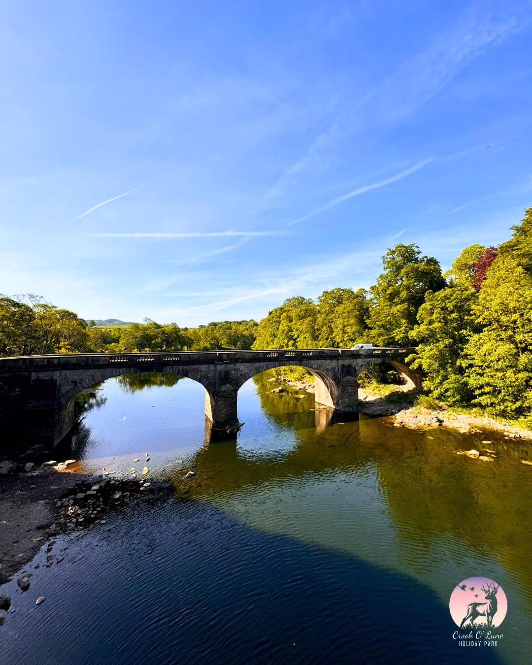 Stone bridge spanning tranquil river with lush green trees and blue sky