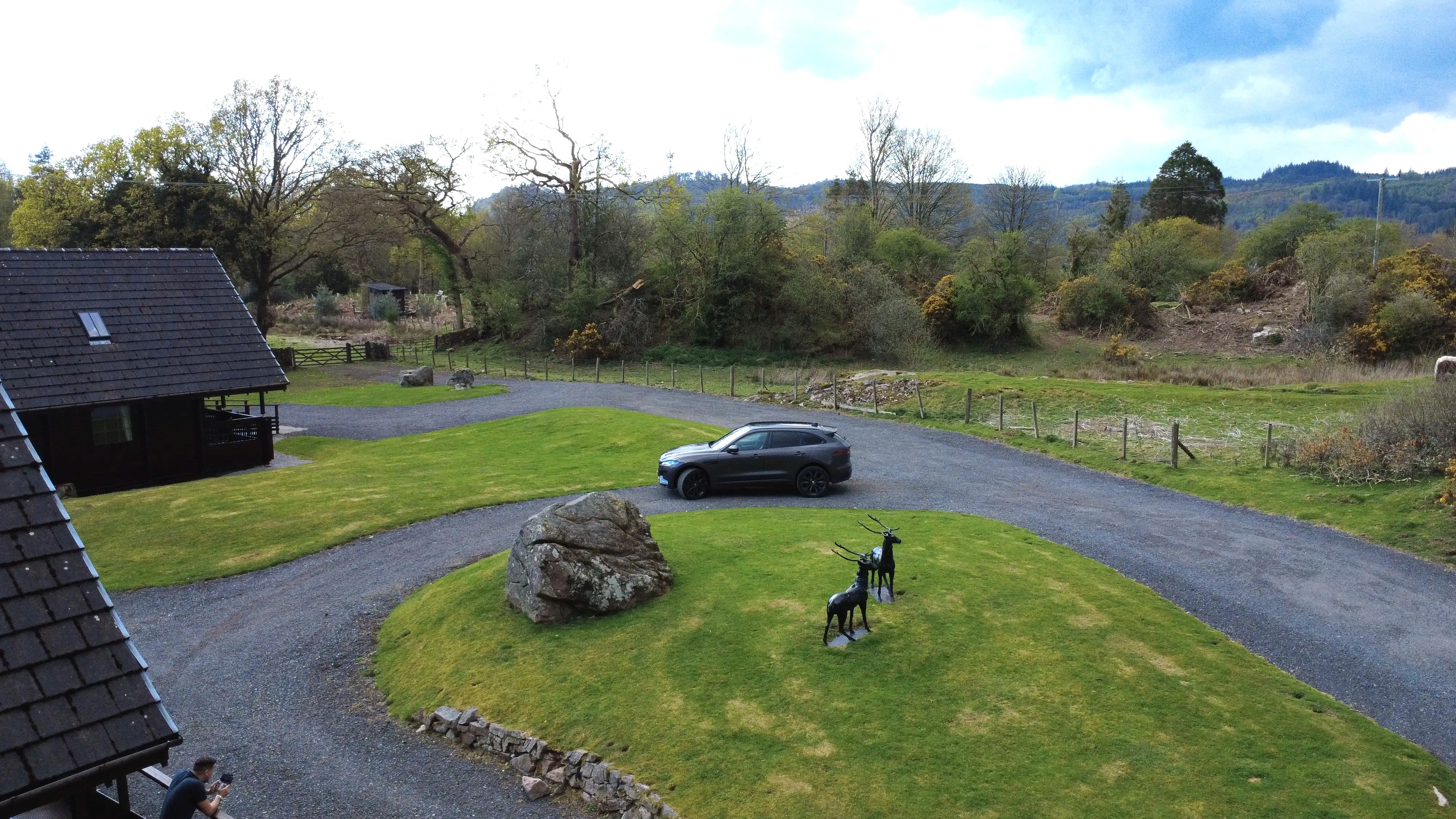 Gray car parked near circular driveway with rock and deer statues in rural landscape