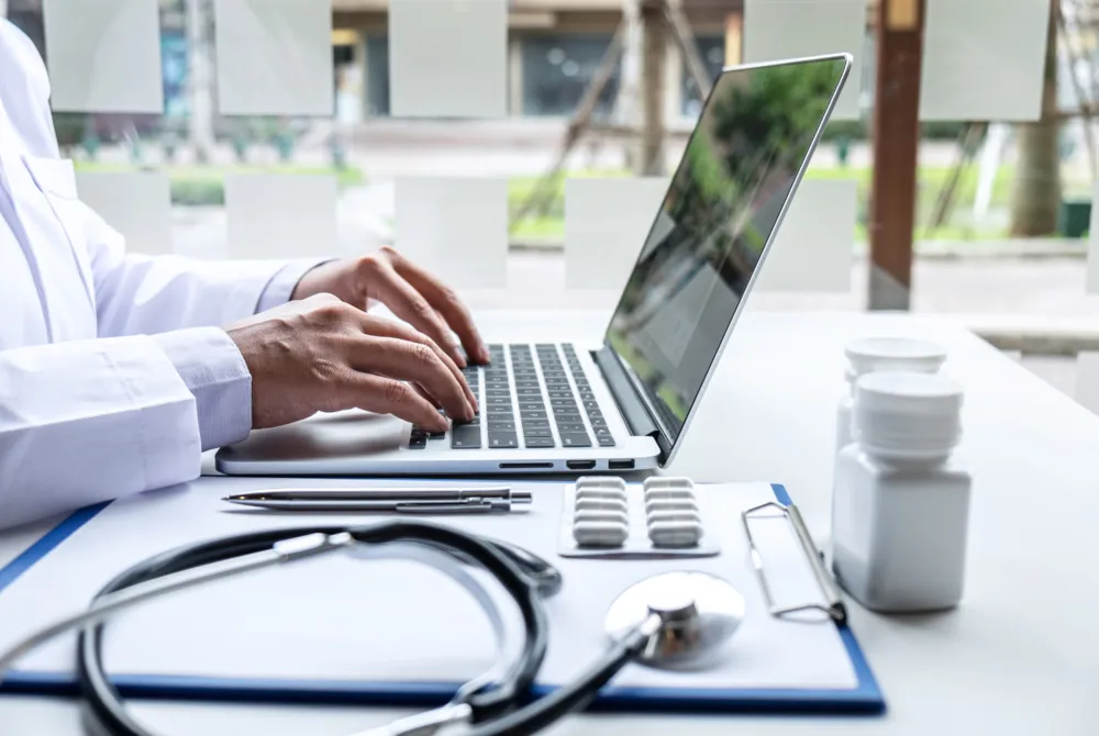 Un professionnel de la santé tape sur un ordinateur portable avec un stéthoscope, des pilules et des bouteilles de médicaments posés sur une table devant lui.