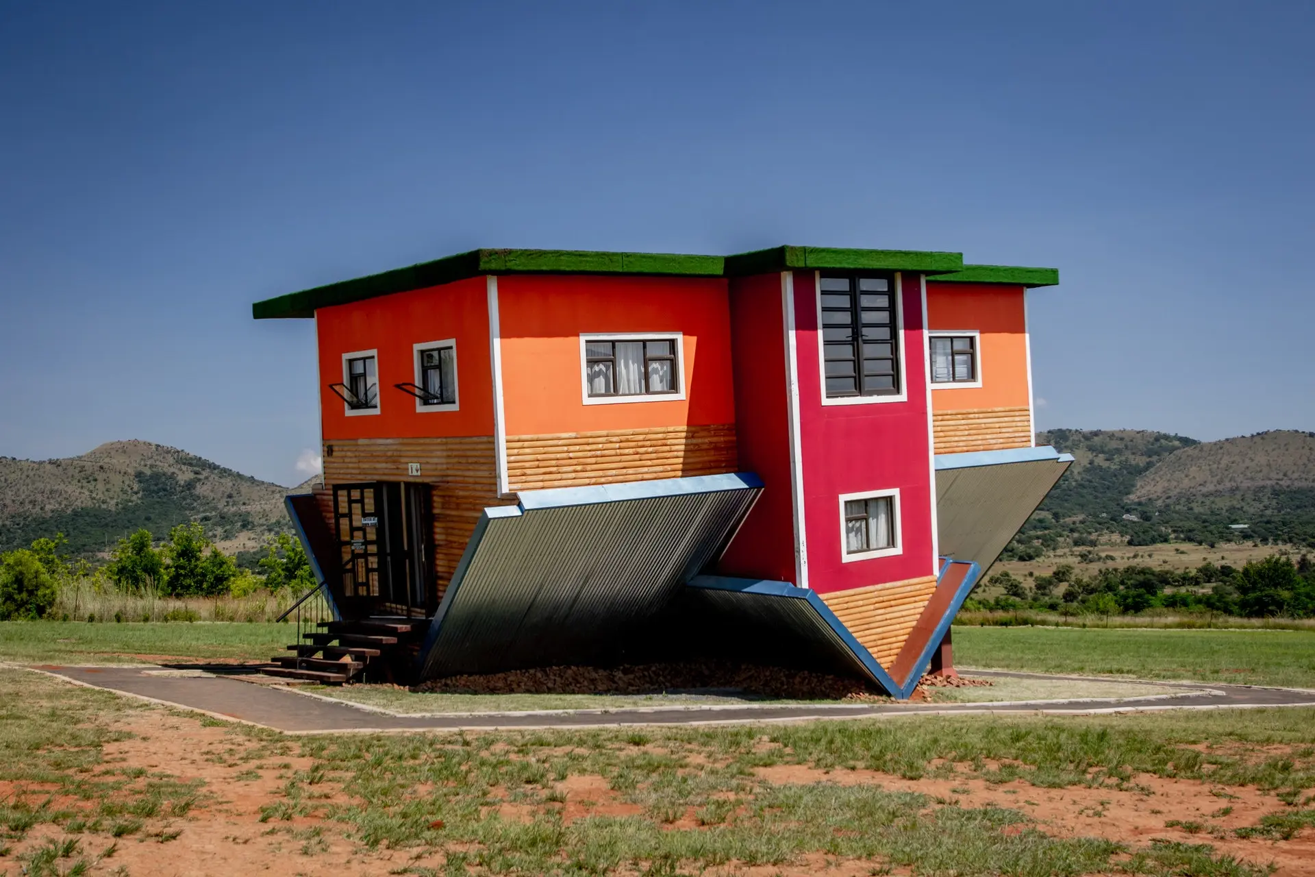 A brightly colored house built upside-down, standing in a field under a blue sky.
