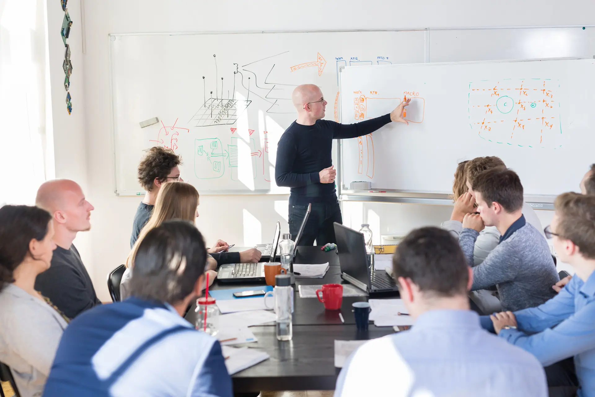 Speaker presenting technical diagrams on a whiteboard to an attentive audience in an office.