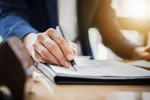 Hand holding a pen, signing a business contract document on a wooden desk.
