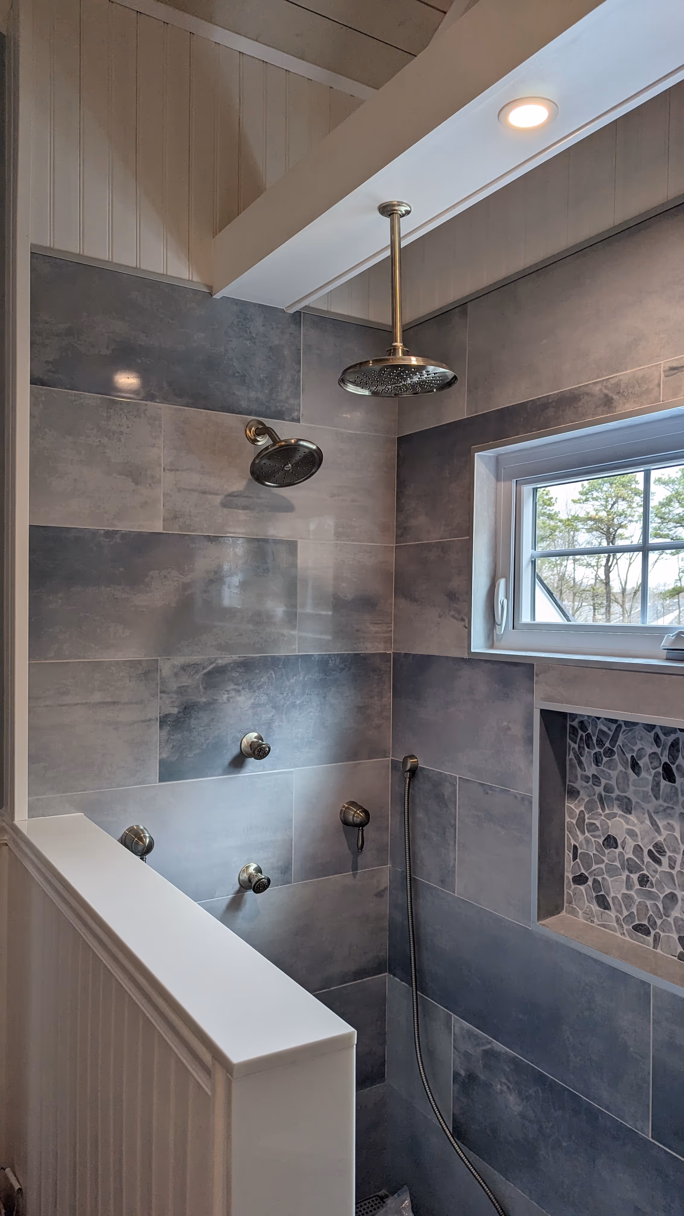 Modern shower area with gray tiles, dual showerheads, a handheld shower, and a small window showing trees outside.