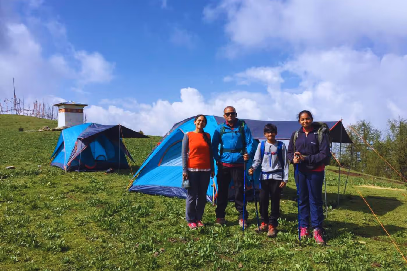 Family of four stands smiling in front of blue dome tents at Bumdra campsite under a bright blue sky, geared up with backpacks and hiking poles.