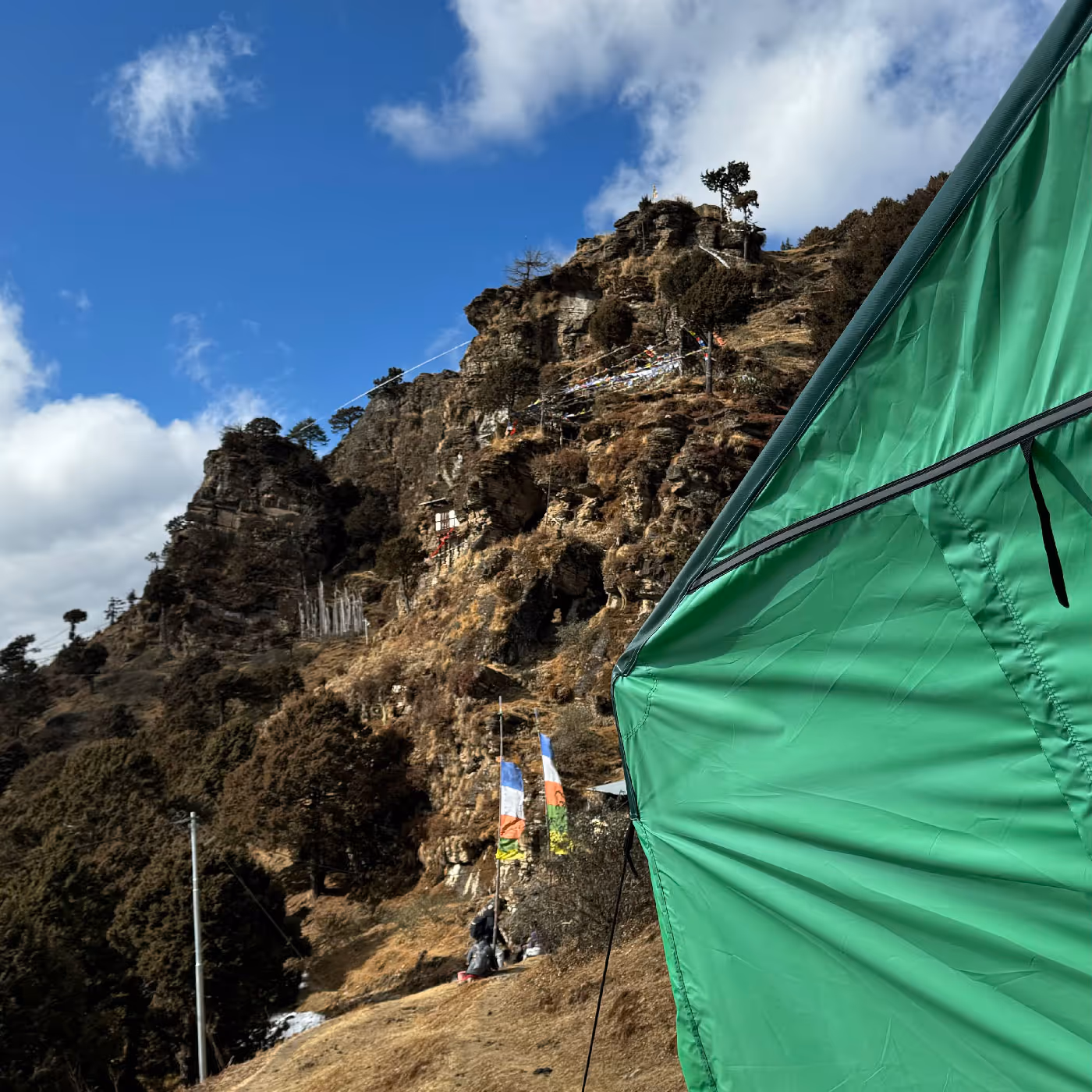 Green trekking tent pitched at Bumdra campsite with towering cliffs and prayer flags fluttering in the background.